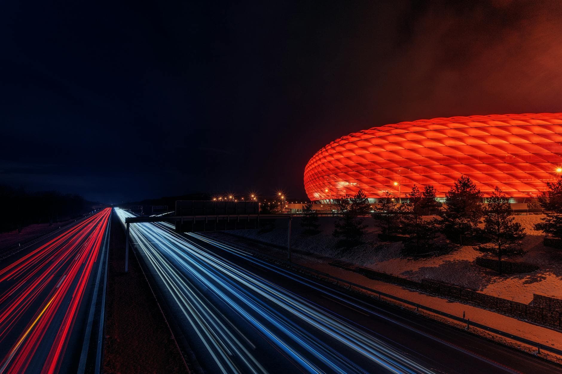 Die nachts rot beleuchtete Allianz Arena in München, davor die Autobahn mit Lichtspuren fahrender Autos